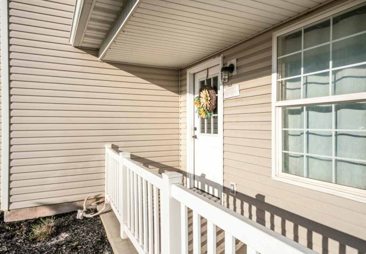 Front door exterior of a house with vinyl wall sidings. There is a white wooden railings at the front of a door with glass panel and wreath beside the double hung window.